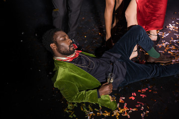 handsome and drunk african american man lying on floor with confetti near friends on black