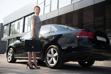 low angle view of happy businesswoman standing near black car with briefcase