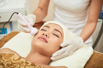 Fototapeta na wymiar Asian beautiful woman lying with eyes closed while cosmetologist doing ultrasound procedure for her face in modern spa salon