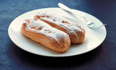 Two eclairs with powdered sugar on a white plate, with a fork, on a dark background.