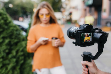 Male photographer working in front of blonde girl in trendy sweater which standing on the street. Outdoor photo of lovely young woman with hand and camera on foreground.