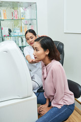 Asian young woman determining her skin type with modern equipment together with her dermatologist in office