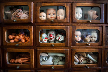 Funny and ugly faces of dolls inside wooden house with small windows. Many parts of heads, hands and legs inside small boxes. Broken toys and details for children games
