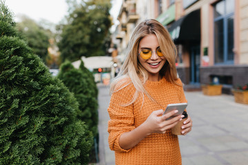 Lovely blonde girl walking by green bushes with smile, carrying smartphone and cup of coffee....