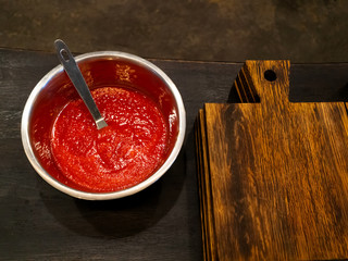 Tomato sauce in a metal bowl on a dark wooden table near a wooden cutting board before cooking pizza
