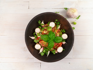 Vegetarian salad with greens and champignons in a dark wooden plate on a wooden table in white. Near the plate is a head of garlic, close up