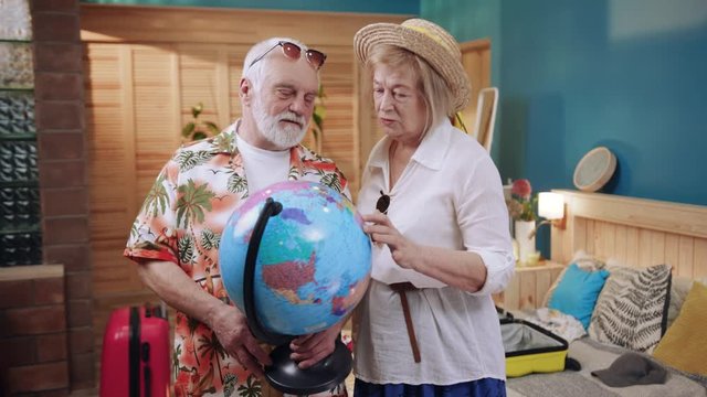 Elderly couple holding globe in hands and choosing new travel destination. Old travelers. Happy ageing. Home. Journey, adventures. Indoors. Pink suitcase in background.
