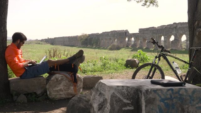 Cyclist with bike sitting on a rock under a tree writing on laptop in front of ancient roman aqueduct. Young attractive athletic man with orange sportswear and backpack in parco degli acquedotti in