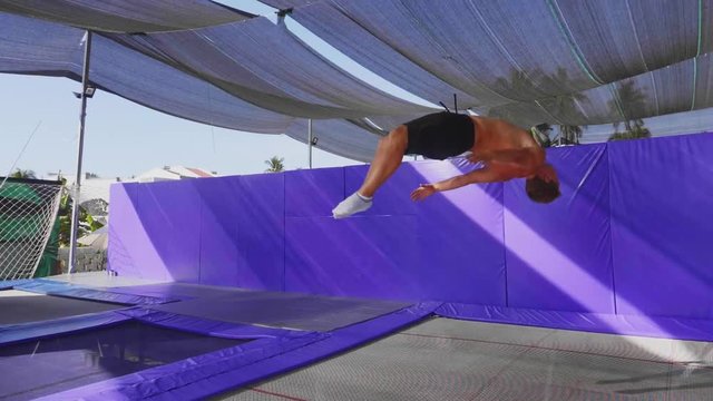 Professional man gymnast with muscular body in black shorts with naked torso is training on trampoline making back somersault outdoor on tropical island background, slow motion.