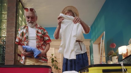 Cheerful elderly Caucasian couple standing near suitcases and packing up things, choosing clothes for trip. Tourism concept. Indoors. Good mood. Blue walls of room. Home. - Powered by Adobe