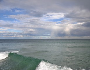 A clear blue sky is visible through the gray clouds on the sea. In the foreground a green wave. In the distance the horizon line.