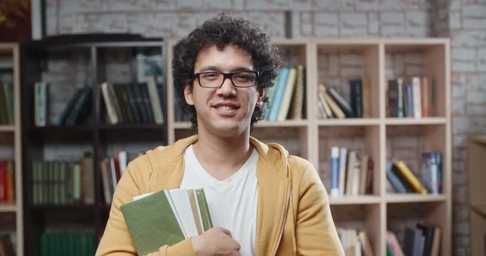 Asian guy with curly hair is standing between bookshelves of library holding books in hands, looking at camera and smoling - portrait closeup 4k, education, student lifestyle concept 4k