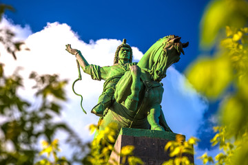 Majestic bronze green monument to Salawat Yulaev, close-up shot through spring foliage on a bright summer day. Folk hero on a horse with whip in his hand. Salavat Yulayev, Ufa, Bashkortostan, Russia.