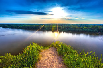 Drowned gravel dangerous path before a cliff overgrown green grass, rise the White River during sunset under a clear blue sky against the setting sun. Rock Hanging Stone, Ufa, Bashkortostan, Russia.