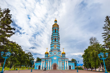 Light blue high church with golden domes surrounded by green trees under large rain clouds on the...