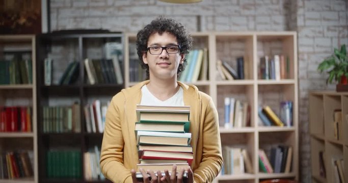 Asian guy with curly hair is standing between bookshelves of library holding books in hands, looking at camera and smoling - portrait closeup 4k, education, student lifestyle concept 4k