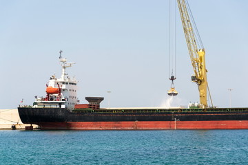 Fototapeta premium Yellow crane unloading sand from large freighter cargo ship in harbor, freight digitalization, transportation efficiency, sand shortage concept, sunny day copy space