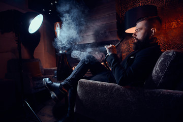 Bearded relaxed man is sitting on the armchair and smoking hookah near fireplace.