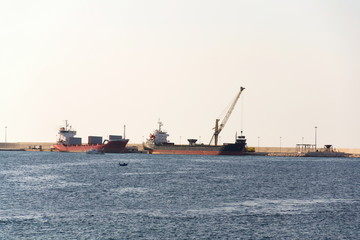 Fototapeta premium Yellow crane unloading sand from large freighter cargo ship in harbor, freight digitalization, transportation efficiency, sand shortage concept, sunny day copy space