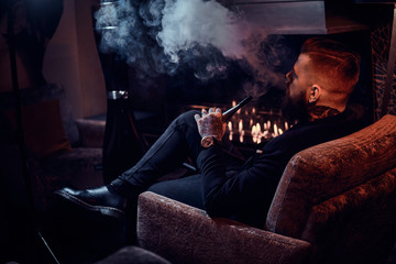 Bearded relaxed man is sitting on the armchair and smoking hookah near fireplace.