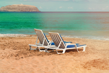 Two empty deck chairs on a deserted sandy beach on the ocean.