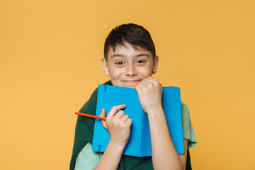 Young cute caucasian boy, wears  green shirt and green sweater, holds blue exercise book and red pencil and happily smiling. models over yellow studio wall. Positive people concept. holidays began