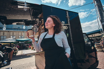 Pretty young barista has a break and enjoying coffee. There are bright sunny summer day and windy. She is wearing uniform.