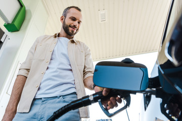 low angle view of happy man holding fuel pump while refueling automobile with benzine at gas station