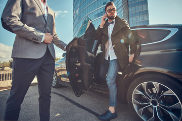 Handsome businessman in sunglasses is going out from car while his assistant is opening door for...
