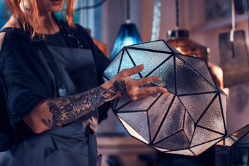 Talanted young designer with tattoo on her hand is prepearing new lamp, she is holdind it in hands.