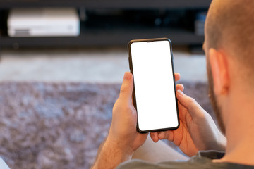 A white Caucasian male holds a black smartphone with a big white screen in a moden day living room with the TV in the background. Phone is isolated and ready for pasting over.