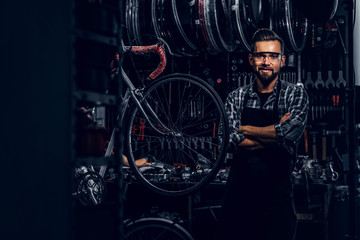 Smiling bearded man in glasses is standing near fixed bicycle at his own workshop.
