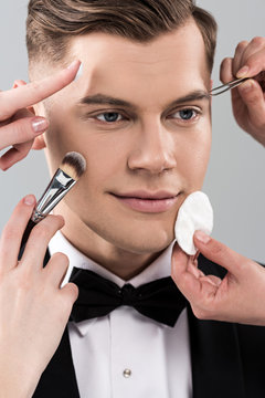 cropped view of makeup artists doing makeup for man in formal wear holding glass of cocktail isolated on grey