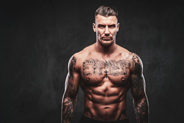 Portrait of a tattooed muscular shirtless man with stylish hair posing at the camera on a dark...