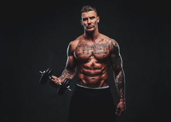 Fototapeta na wymiar A tattooed muscular man doing exercises with dumbbells for biceps on dark background.
