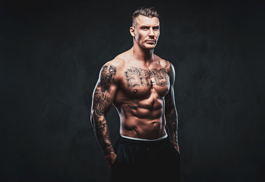 A tattooed muscular shirtless man with stylish hair posing at the camera on a dark background.