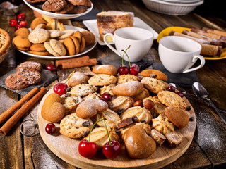 Oatmeal cookies and sand chocolate cake with cherry on cutting board on wooden table in rustic style served with cups and dessert spoons. Breakfast for loved one.