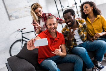 Selective focus of cheerful multicultural friends taking selfie on smartphone at home