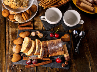 Oatmeal and nut cookies heart shape and sand rolled biscuit on tier cake stand and cinnamon stick on kitchen on wood table in rustic style. Cups of tea and dessert spoon. Top view.