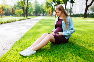 Naklejka na ściany i meble Happy young pregnant woman sitting in the park on the lawn,