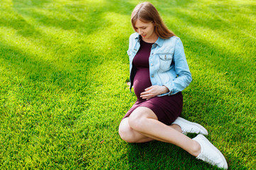 Fototapeta premium Happy young pregnant woman sitting in the park on the lawn,