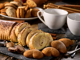 Sand cookies heart shape, rolled cake with cherry and cinnamon stick, coffee cups on kitchen on wooden table in village style. Useful for digestion. Limitations for diabetics.