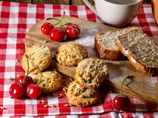 Oatmeal cookies with husk oats and cherry cupcake with powdered sugar on kitchen cutting board and cup of tea gingham checkered cotton fabric on table in village style for picnic. Coffee in cafe.