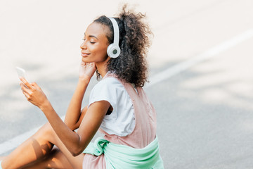 smiling african american girl listening music in headphones and using smartphone on road
