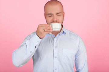 Portrait of a beautiful bald man with a beard dressed in a white shirt. sniffs coffee from a cup that has risen to the nose. stands in front of the pink background