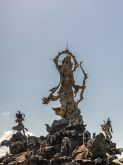 Denpasar, Bali, Indonesia - February 26, 2019: Closeup of Patung Titi Banda brown stone statue of holy warrior in center of roundabout of By Pass Ngurah Rai Street under light blue sky.