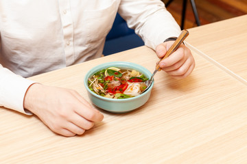 Man is having spicy thai noodle soup, served on a wooden table, for lunch in a cafe