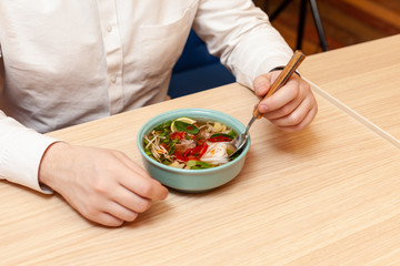 Man is having spicy thai noodle soup, served on a wooden table, for lunch in a cafe