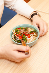 Man is having spicy thai noodle soup, served on a wooden table, for lunch in a cafe