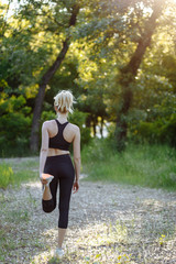 Woman stretching in the summer park before fitness training session at sunset. Young blonde girl warming up outdoors. She is stretching her legs before run